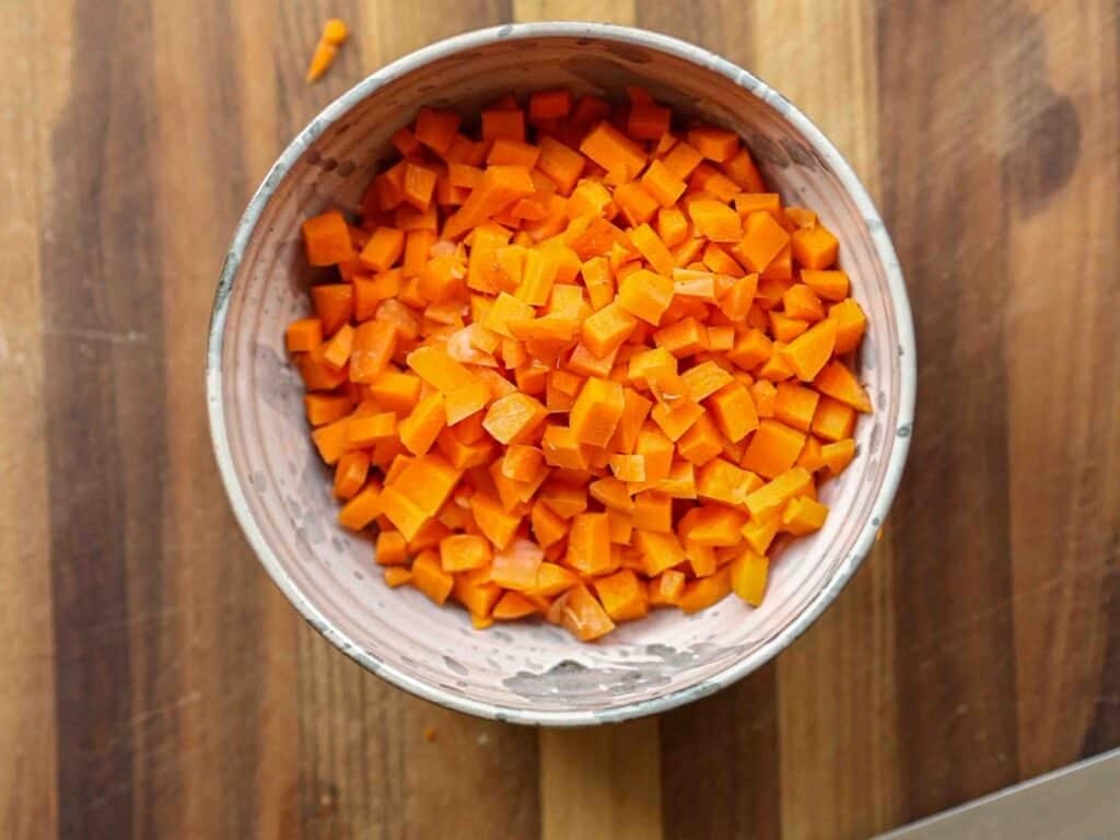 A bowl filled with small, evenly diced orange carrots sits on a wooden surface with visible grain patterns.